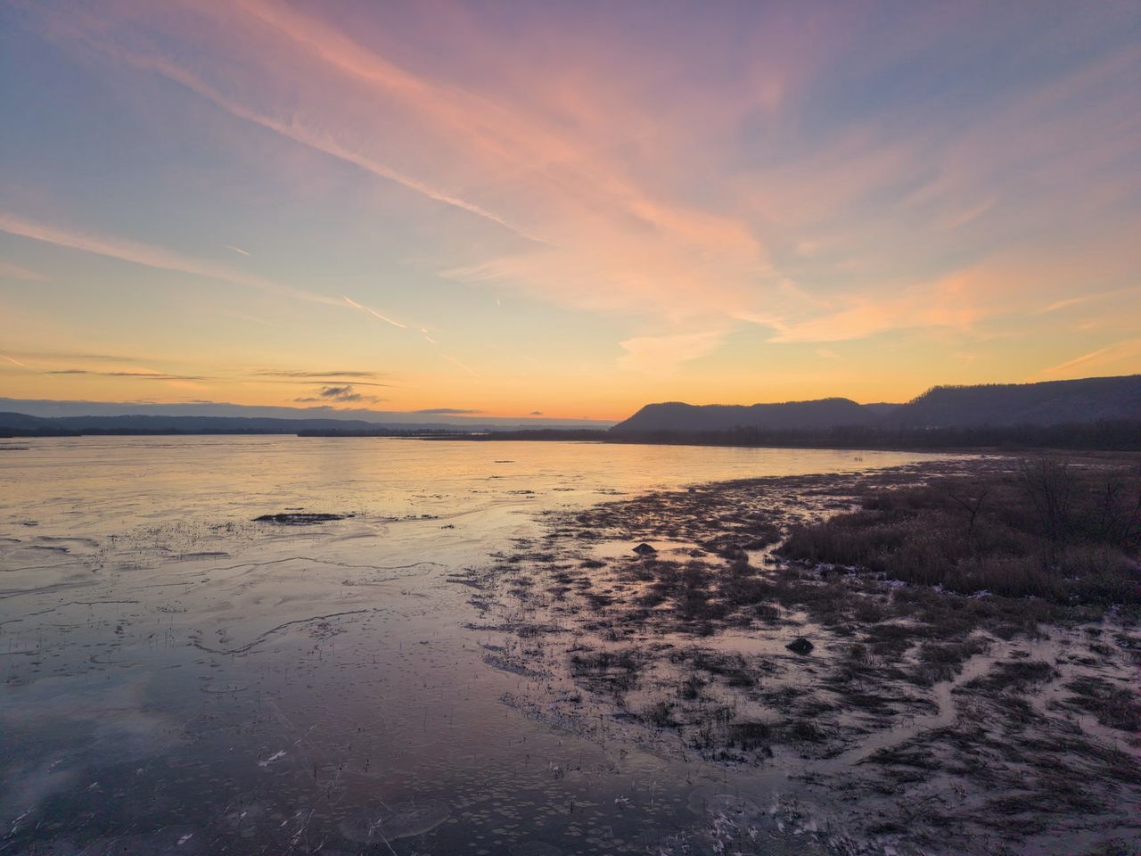 Estuaire gelé au crépuscule, ciel rose et orangé au-dessus d'une rive de marais