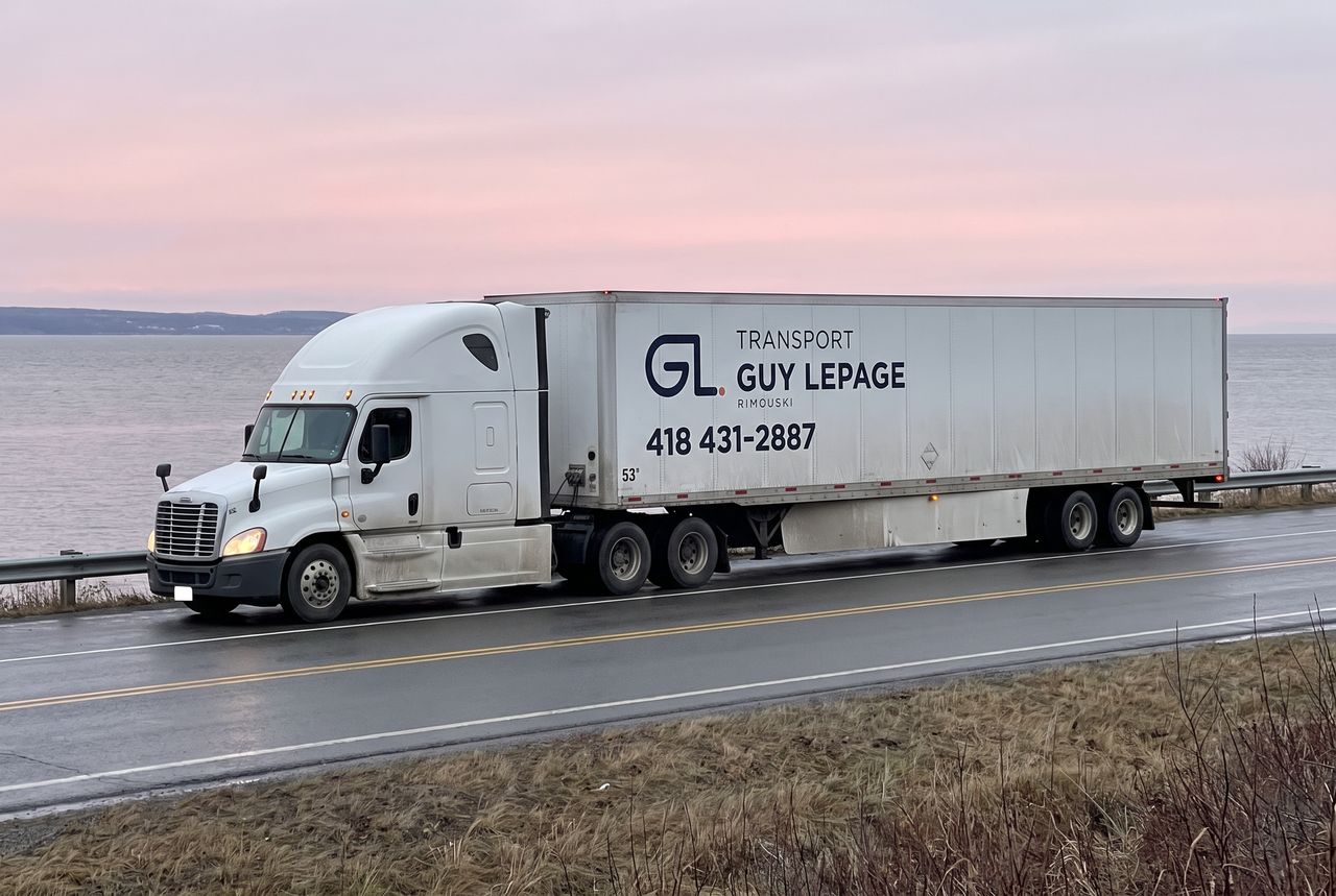 Tracteur routier blanc et remorque aux couleurs de Transport Guy Lepage, sur la route côtière devant l'estuaire, au lever du jour
