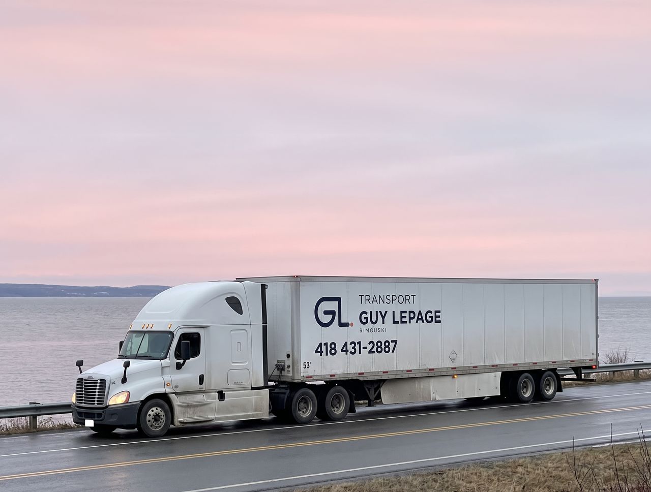 Tracteur routier blanc et remorque aux couleurs de Transport Guy Lepage sur la route côtière, devant l'estuaire, au lever du jour